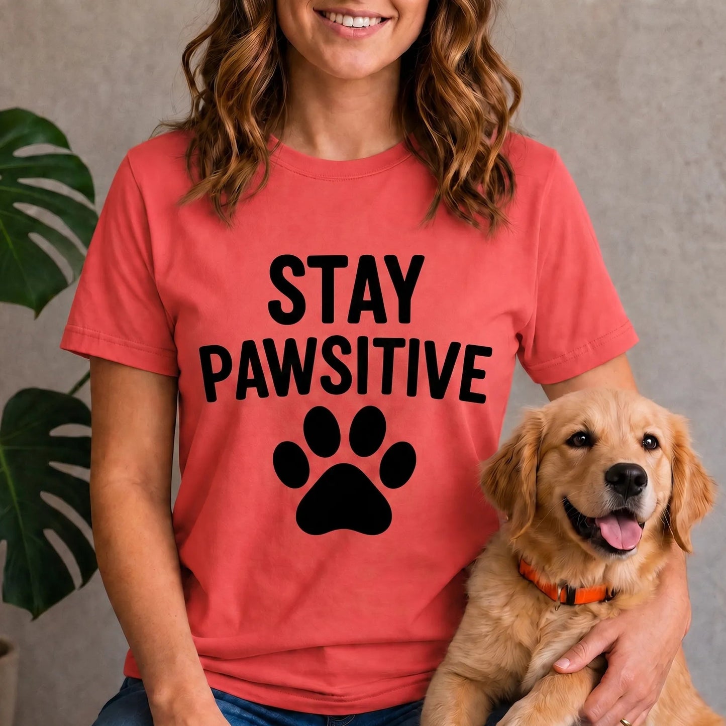 A woman wearing a coral red t-shirt with the black text 'STAY PAWSITIVE' and a paw print graphic, sitting with a golden retriever dog. A potted green plant is visible in the background against a textured gray wall.